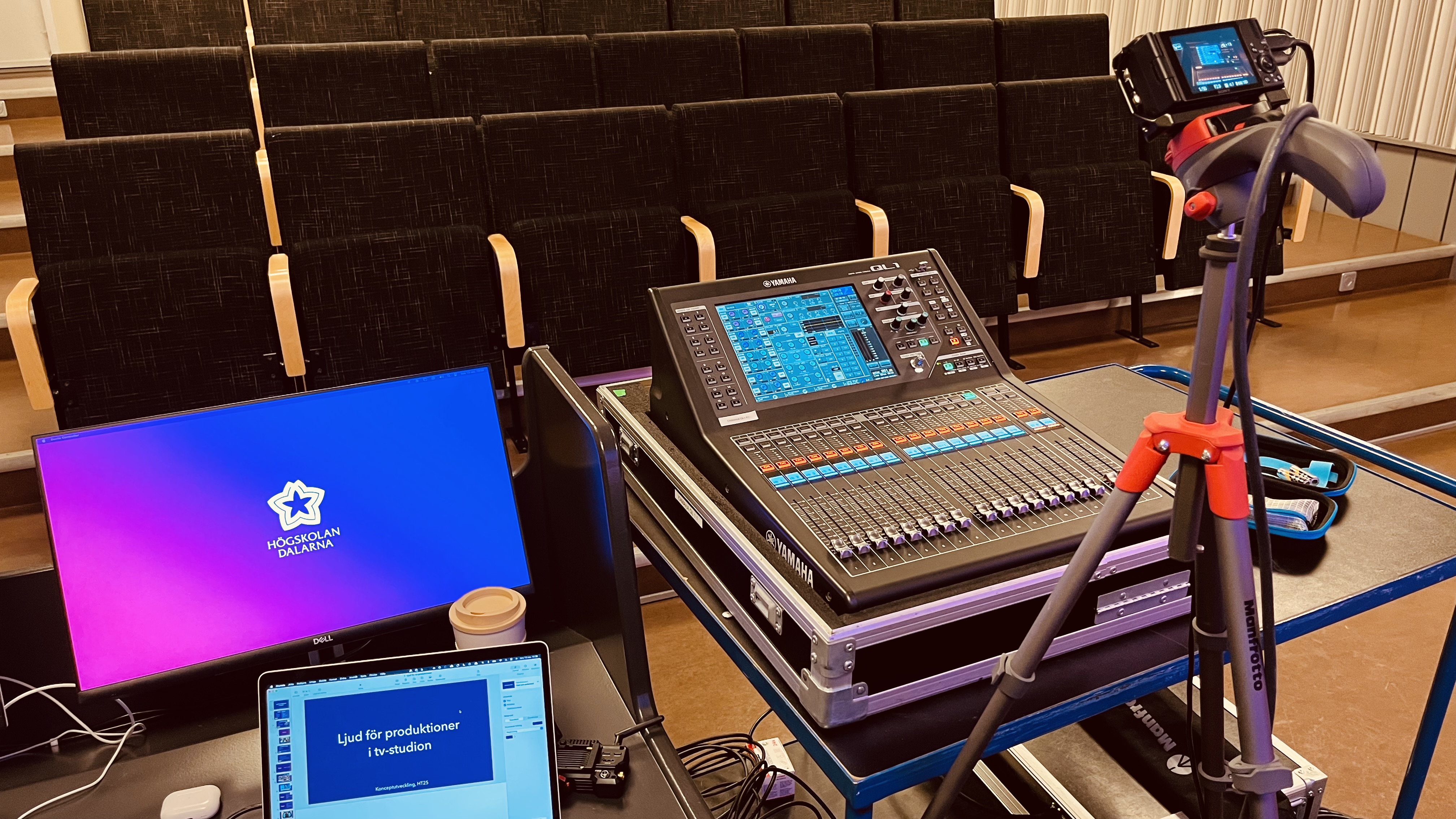 A photo of a lecture room. There's a digital mixing console, a laptop computer and an external display in the center of the image.